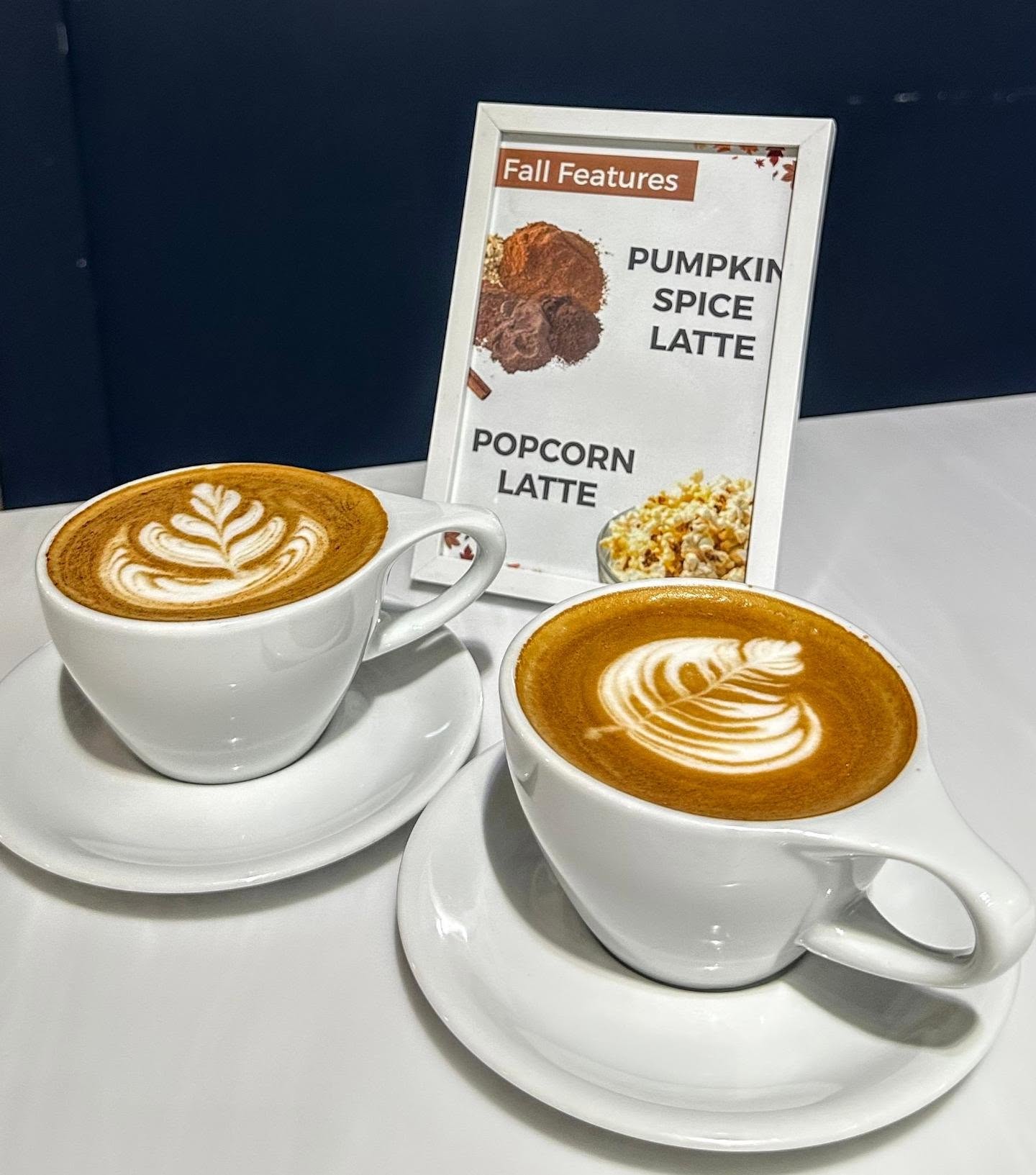 Two lattes with leaf-pattern latte art next to a Denim Coffee Fall Features menu sign