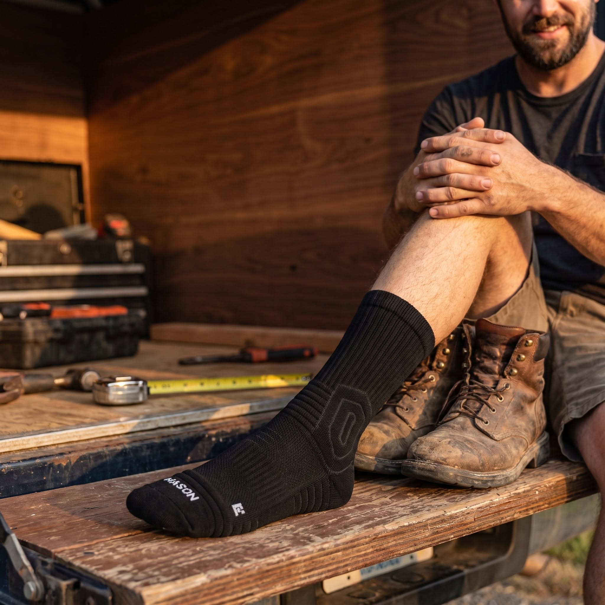 Tradesman sitting on a truck tailgate putting on a Mason sock with work boots nearby