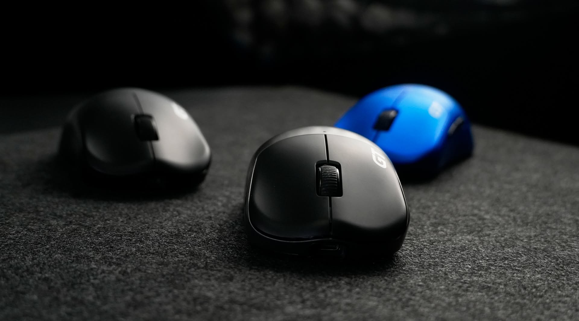 Three RAWM gaming mice in black, grey and blue on a dark mousepad